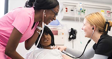 Two nursing students use stethoscopes to monitor heart rate on a model patient.