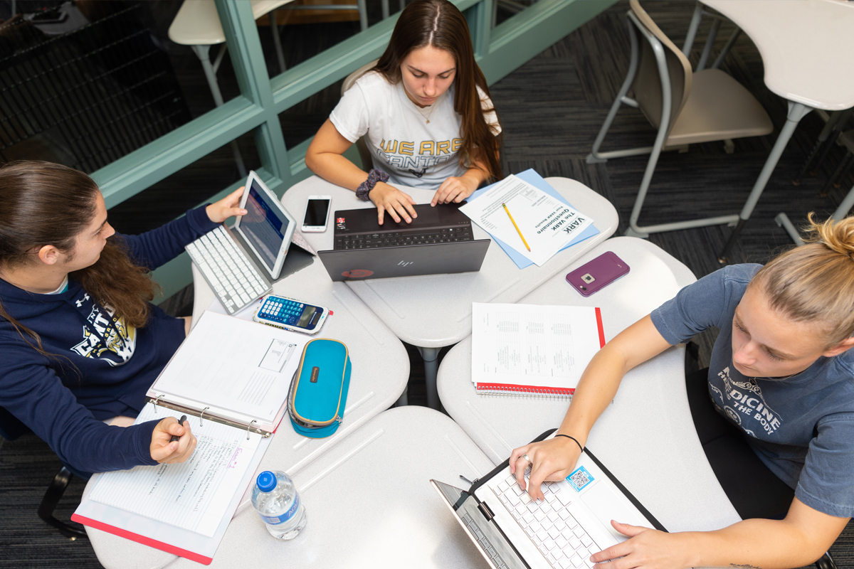Three students study with laptop and notebooks in the library.
