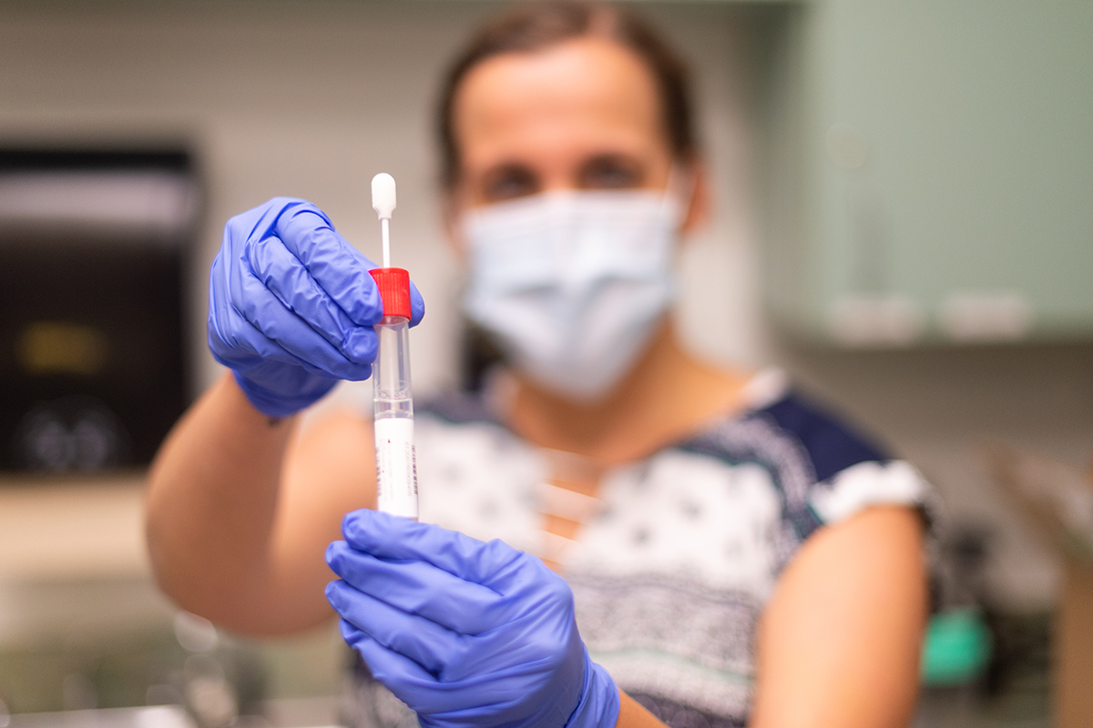 A healthcare worker wearing a mask holds up a COVID-19 testing swab.