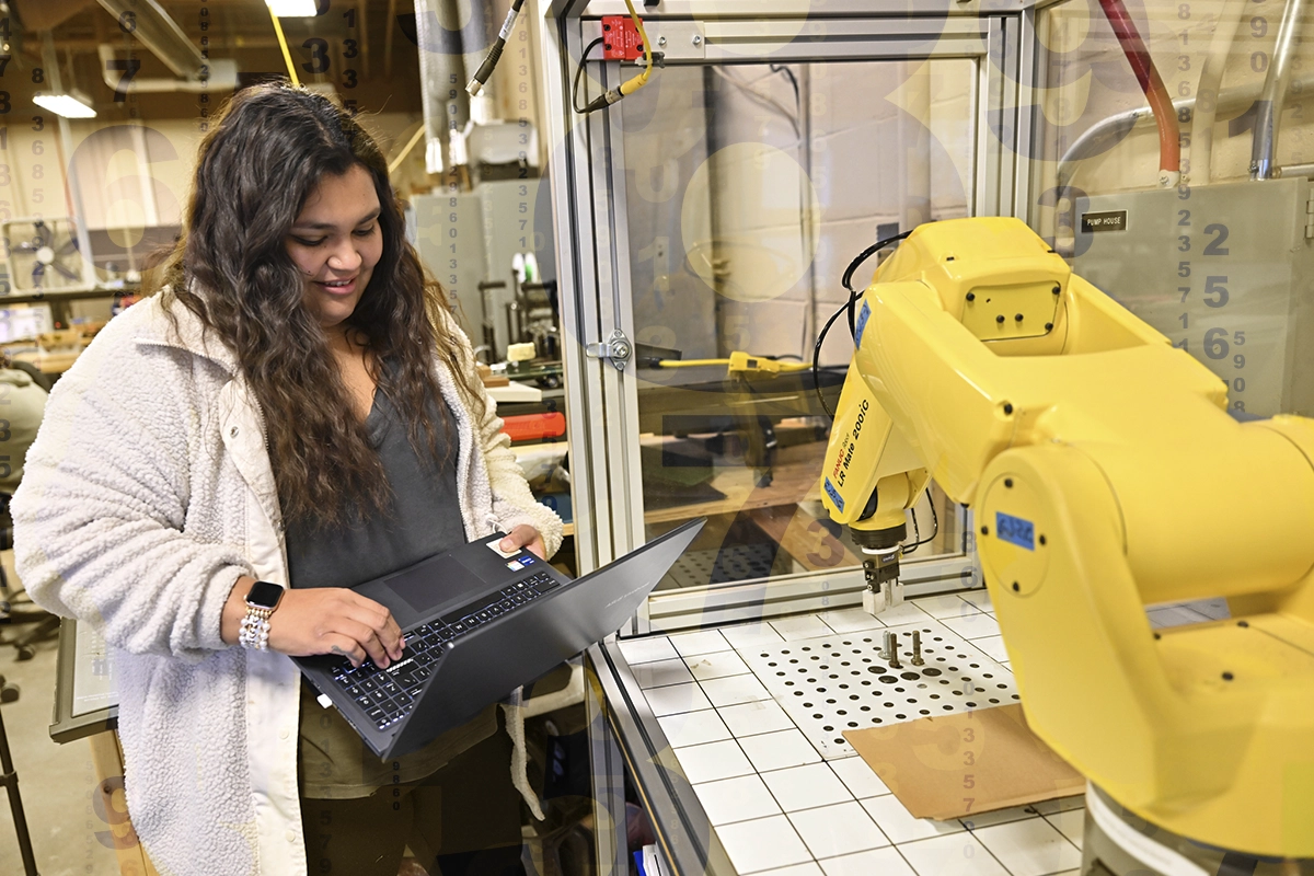 A student uses a laptop to control a mechanical yellow production arm in the Mechanical Engineering Lab.
