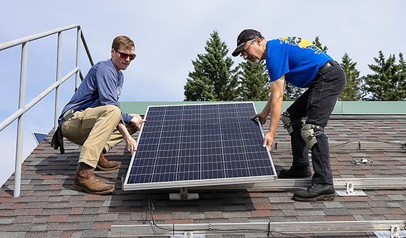 Two students install a solar panel on a mock roof.