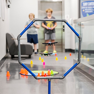 Two campers fly mini drones through a blue hoop during summer camp.