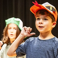 Two campers sing on stage with frog and snake hats.