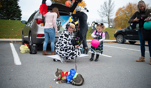 A student dressed as a cow holds a cat dressed as Wonder Woman while a child approaches in a Vampirina outfit.